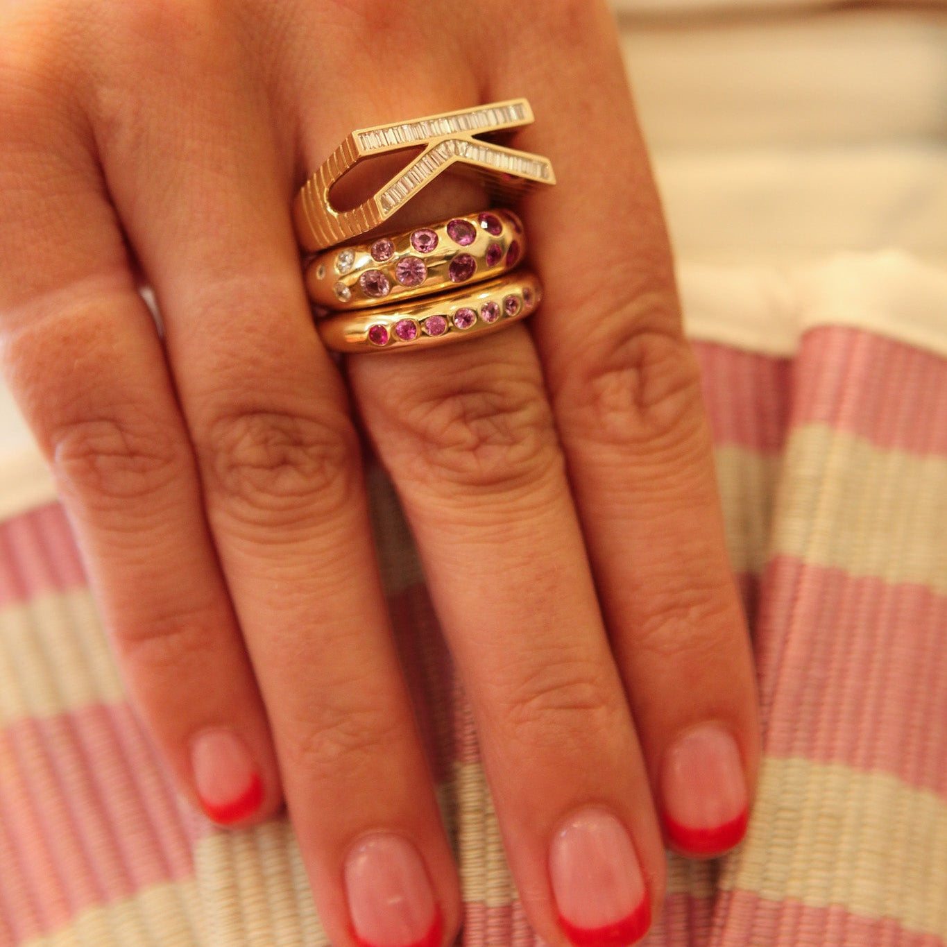 A close-up of a hand wearing three gold rings, including a bow-shaped ring with clear stones and two stacked rings with purple gemstones, resting on a pink and cream striped fabric.