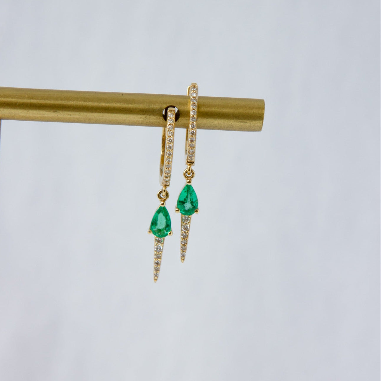 A pair of gold huggie drop earrings with small pavé-set clear stones and a dangling green teardrop gemstone, displayed on a gold stand against a light gray background.