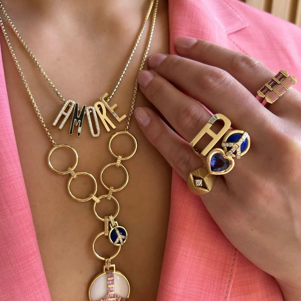 Gold jewelry with 'AMORE' necklace and rings on a pink background