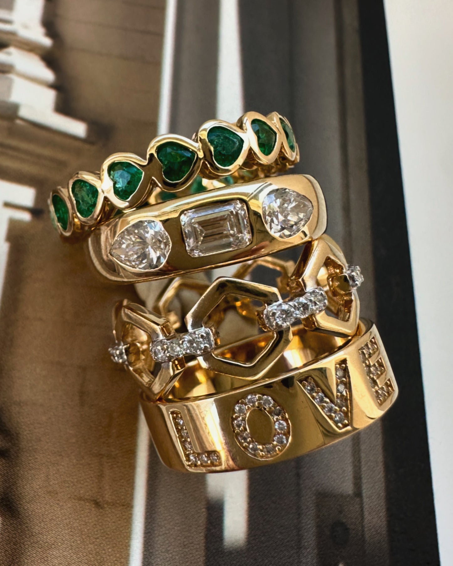 Stack of gold rings with emeralds and diamonds on a blurred architectural background
