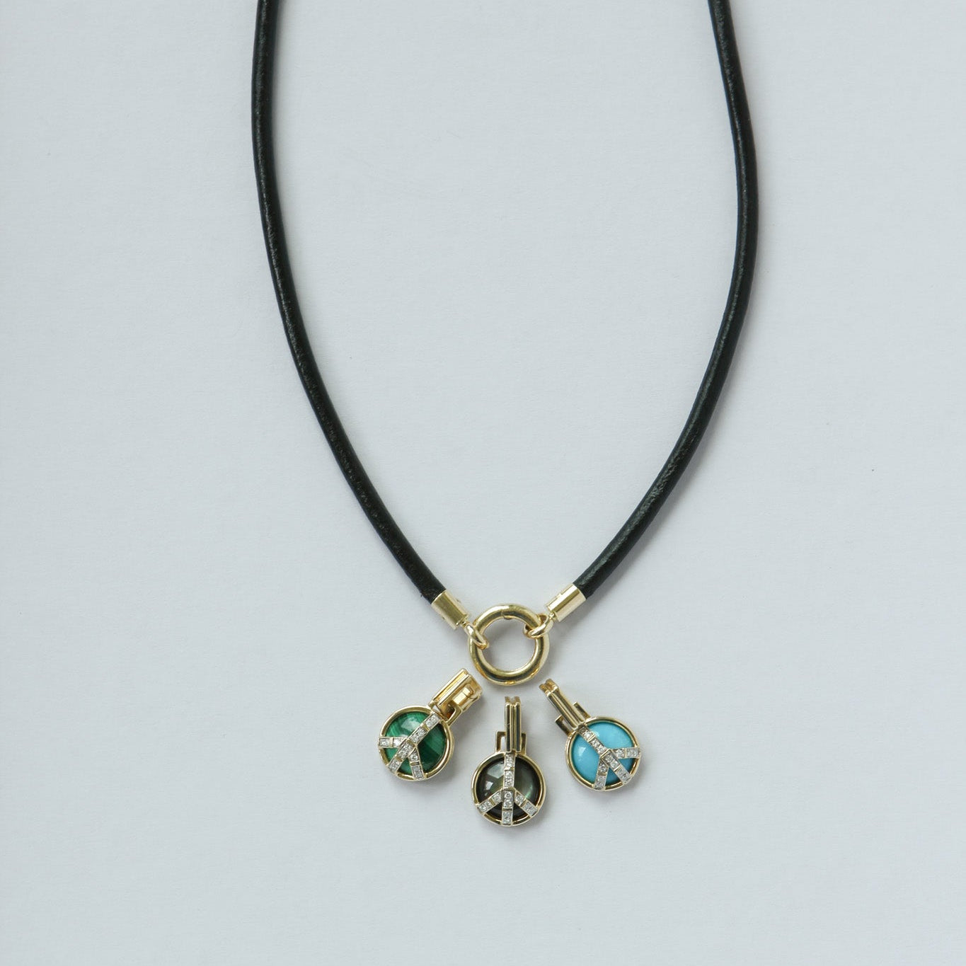 A black cord necklace with a gold-tone ring clasp and three dangling charms: a green enamel peace sign, a turquoise stone charm, and a silver-tone peace sign charm, displayed on a light gray background.