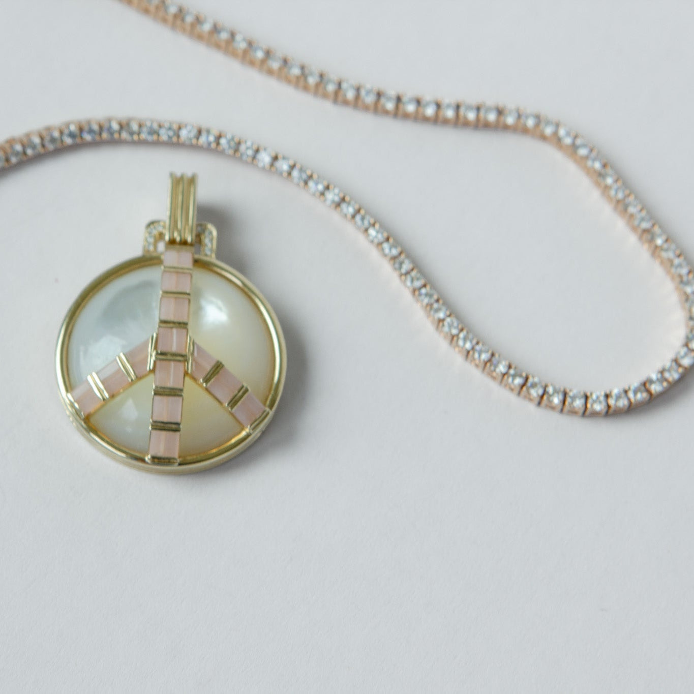 A gold peace sign pendant necklace featuring a milky white mother-of-pearl center, displayed on a delicate gold chain with clear rhinestones against a light gray background.