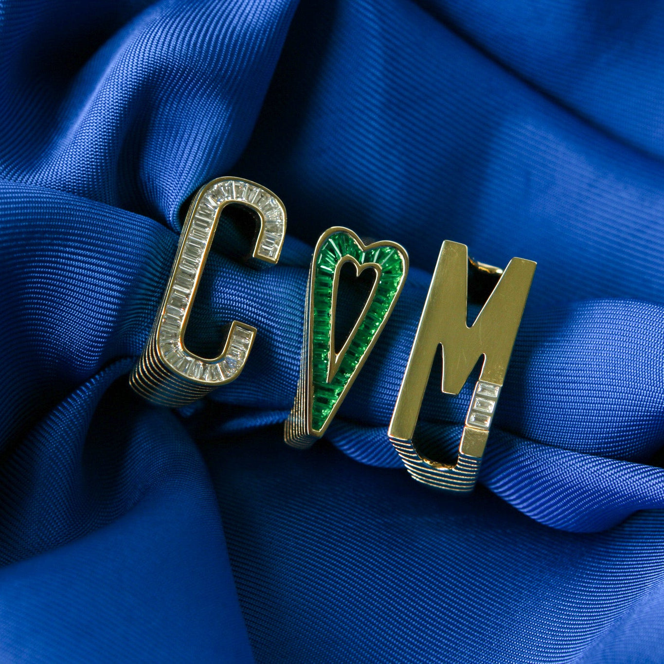 A close-up of a gold-tone ring featuring the letters 'C', 'M', and 'M' arranged to spell 'CMM', with the center letter shaped like a heart and inlaid with green stones. The ring is displayed on draped royal blue satin fabric.