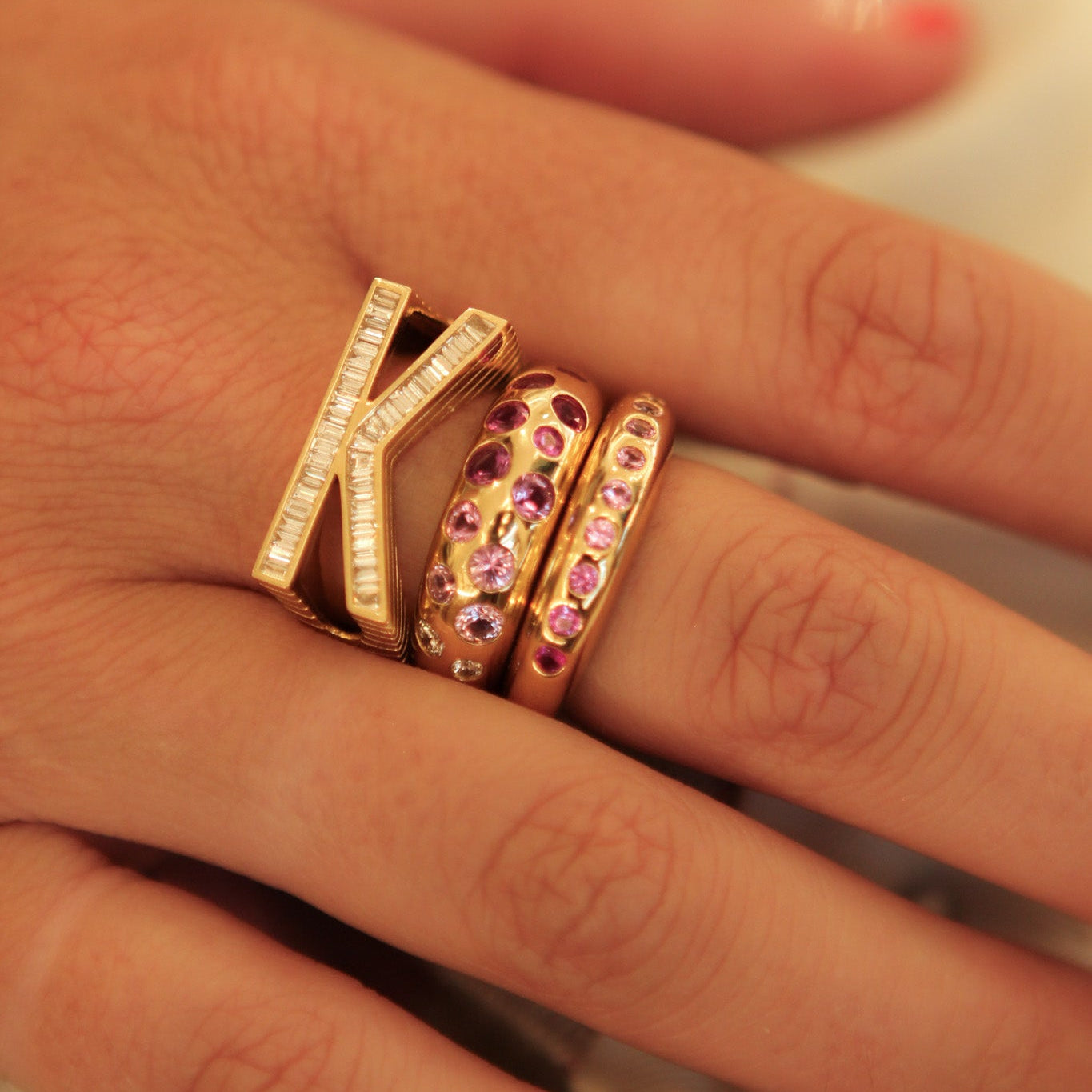 A close-up of a hand wearing three gold rings, including a large letter K ring with clear stones and two smaller rings with pink gemstones.