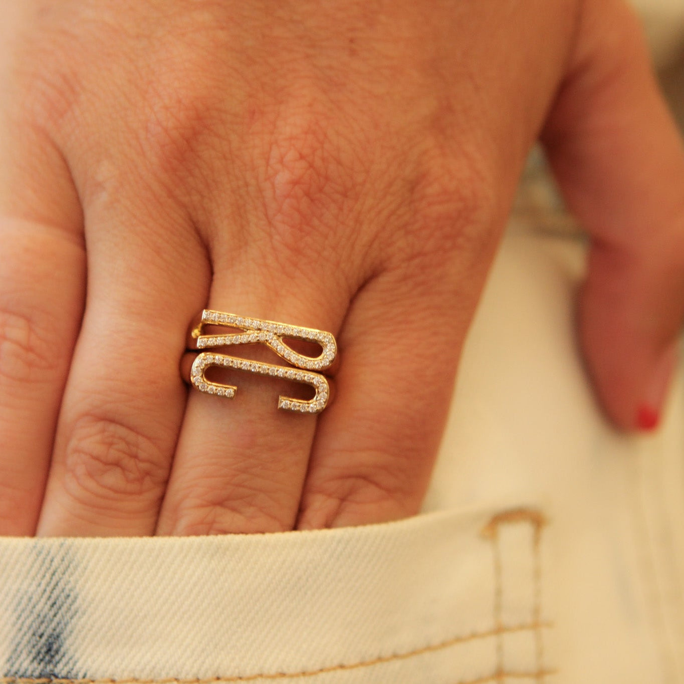 A close-up of a person's hand wearing a gold ring with two intertwined letters 'R' and 'G' set with small clear stones, resting against a light-colored denim jacket.