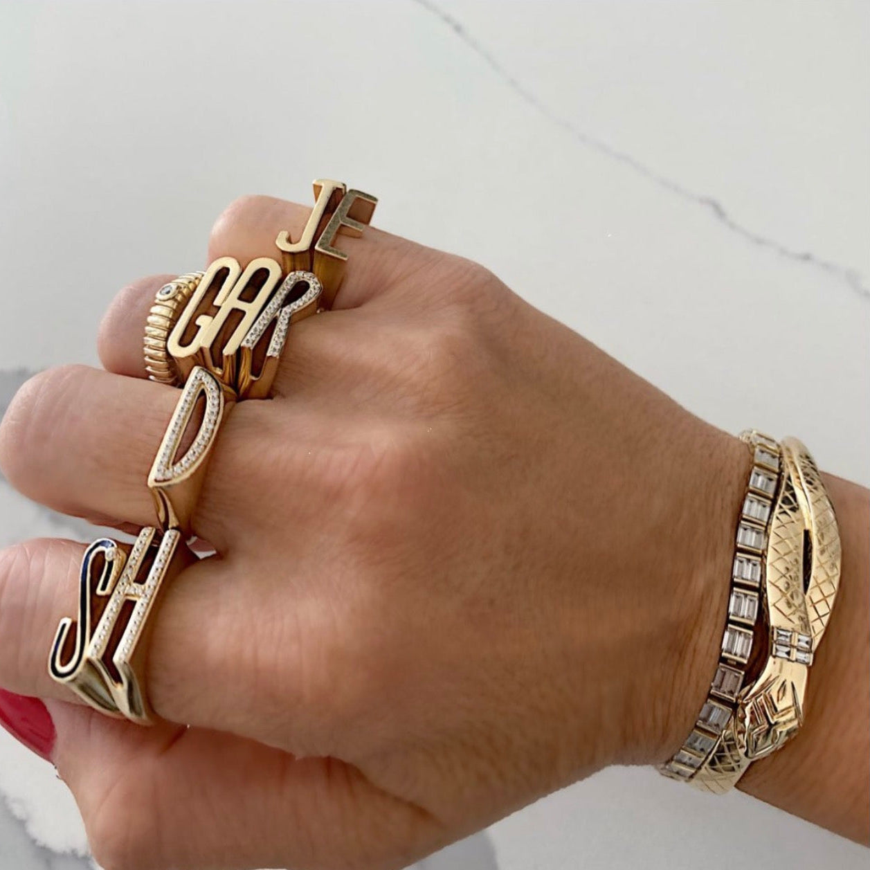 A close-up of a hand wearing multiple gold statement rings and a matching gold bracelet with clear stones, set against a light marble background.