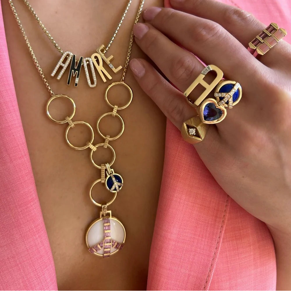 Gold jewelry with 'AMORE' necklace and rings on a pink background