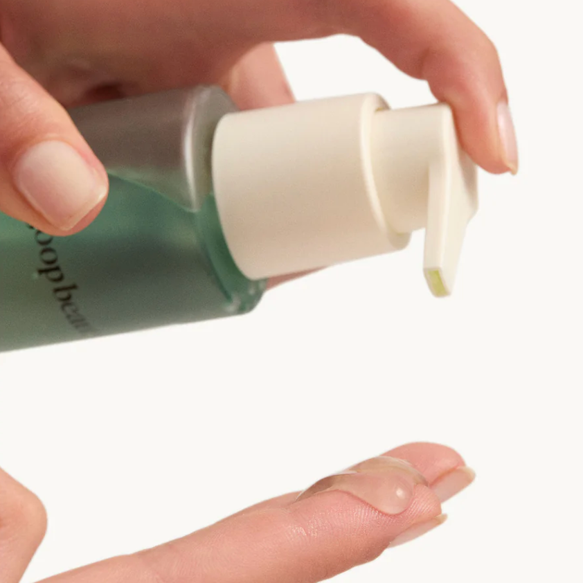 Hand holding a green bottle with a white pump, applying cream to a finger against a white background